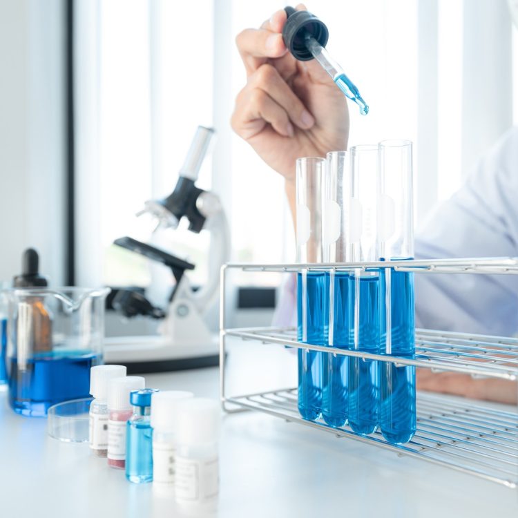 Scientist woman in medical face mask is using dropper to dropping solution into test tube and looking reaction for experimental coronavirus vaccine while working with microscope in laboratory